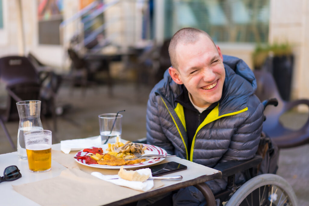 An image of a man sitting in a wheelchair. There is a plate of food on the table along with several glasses of drinks.