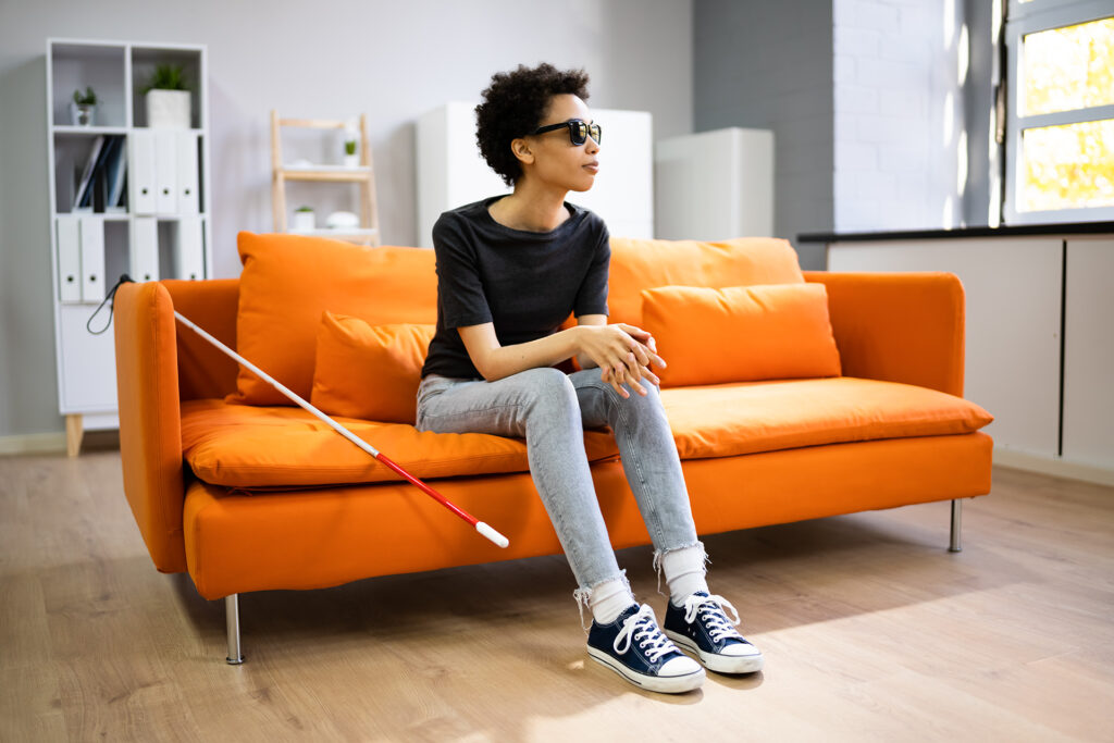 Image of a hotel room with white background and a bright orange couch. A woman of color, wearing dark glasses is sitting on the couch. A guide cane is laying across the couch next to the woman.